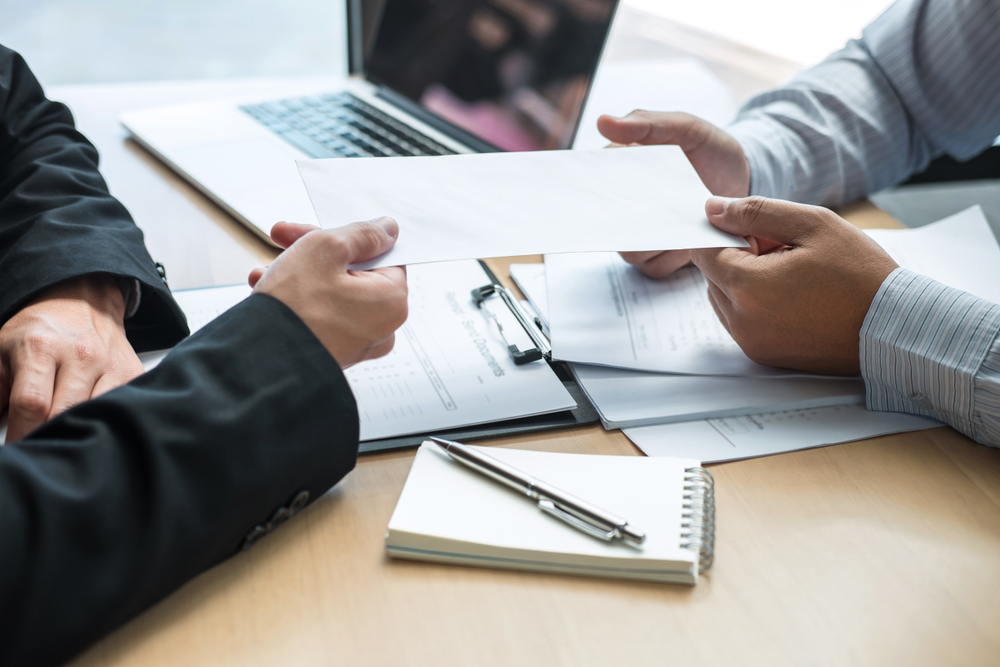 Two business people exchanging a white envelope across a desk with documents and a laptop in the background.