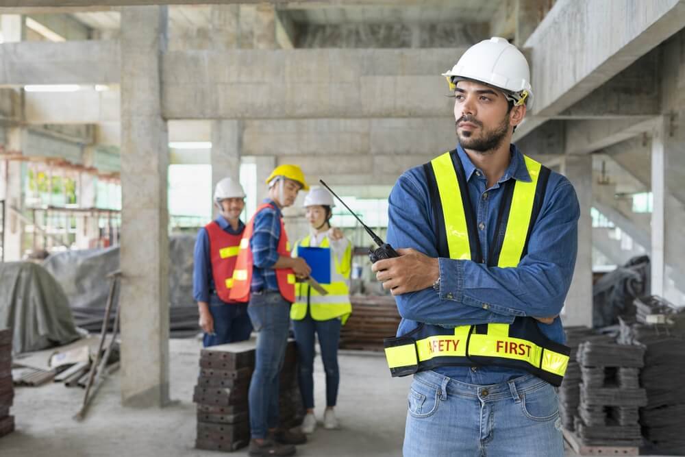 A construction worker wearing a hard hat and safety vest holds a walkie-talkie, while a group of workers in the background discuss something on a clipboard