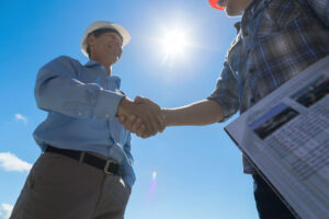 Two men in hard hats shaking hands outdoors under bright sunlight; one holds a document with architectural images, symbolizing a successful agreement or partnership