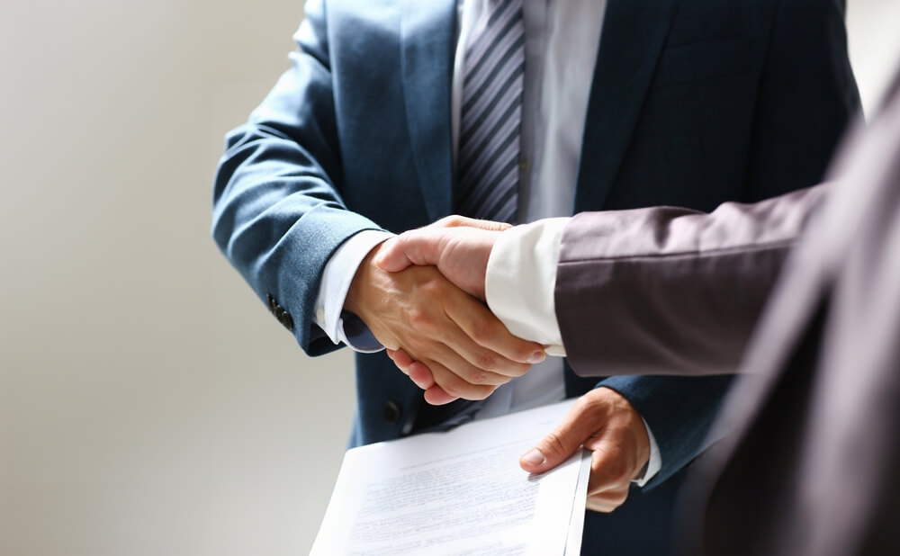 Two people in business suits shaking hands while one holds a document, suggesting the conclusion of a business deal or agreement