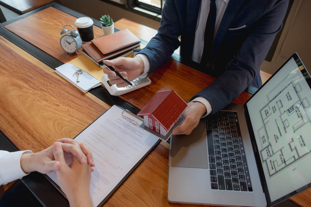 Two people seated at a desk during a real estate meeting; one holds a small house model while pointing with a pen, with a contract, laptop displaying floor plans, and keys on the desk