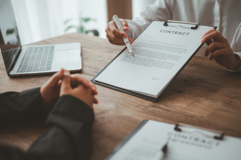 Person in business attire holds a clipboard with a contract, pointing to it with a pen while another person sits across the table with clasped hands. A laptop is nearby