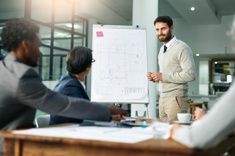 Man presenting a building blueprint to colleagues during a meeting in a modern office. He stands beside a flipchart, engaging the group with his explanation