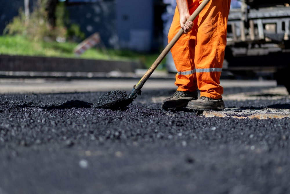 Trabajador con uniforme naranja extiende asfalto fresco con una pala sobre una calle en reparación