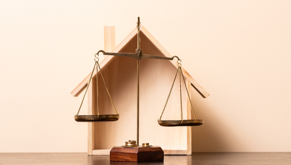 Brass balance scale in front of a wooden house model, symbolizing justice or legal issues in real estate