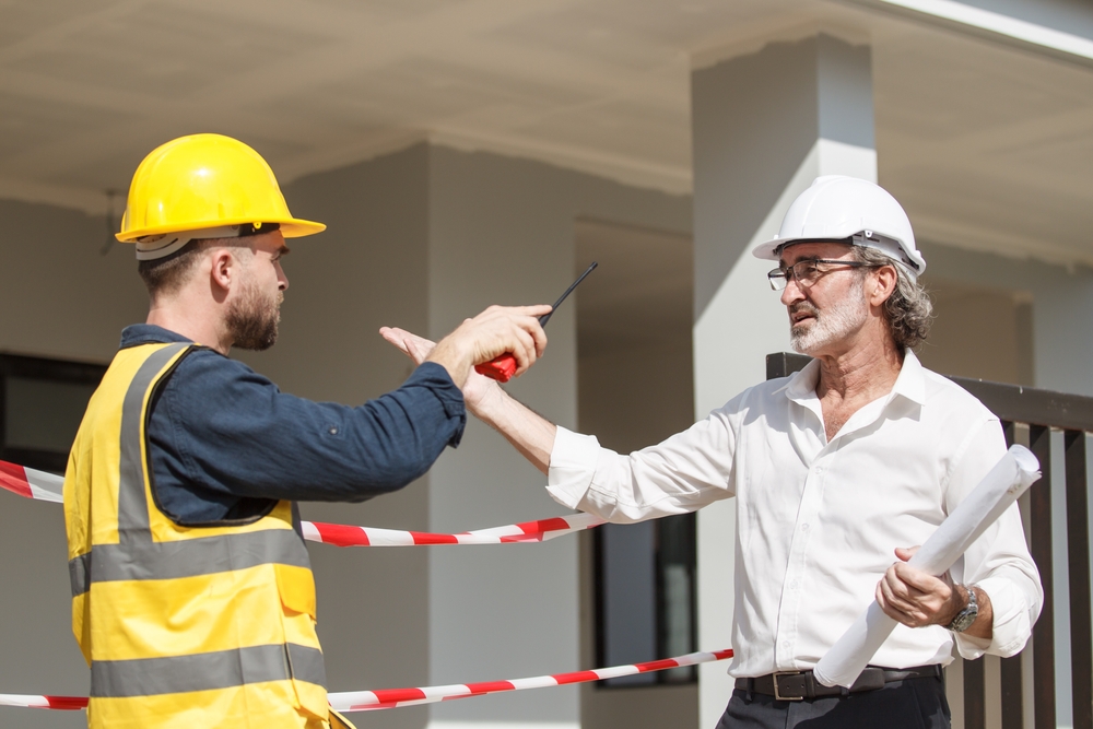 Two construction workers in hard hats discuss work at a building site, gesturing while holding plans